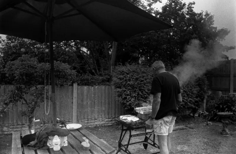 A man grills in a backyard barbecue scene under a patio umbrella