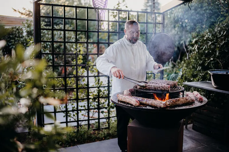 A chef skillfully grills meat on an open flame in a beautiful garden setting, surrounded by greenery
